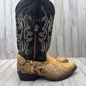 May include: A western-style boot featuring a black leather shaft with intricate gold and silver embroidery and a silver star emblem. The lower portion of the boot is crafted from a textured, yellow and brown snakeskin-patterned material.