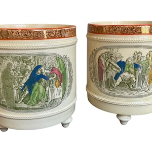 May include: Two white ceramic planters with orange and gold trim. Each planter features a different scene with people in a forest setting. The planters have three legs each.