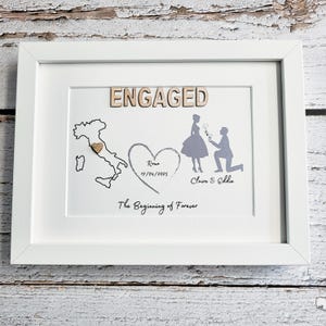 May include: A white framed wall art piece featuring the word ENGAGED in wooden letters, a map of Italy with a small heart, a heart shape containing the text Rome 17/04/2025, a silhouette of a proposal, the names Claire & Eddie, and The Beginning of Forever.