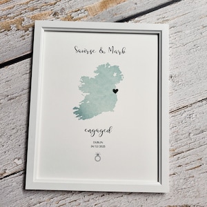 May include: A white framed print featuring a sage green watercolour map of Ireland. The text reads Saoirse & Mark, engaged, Dublin, and 24/12/2025. A small black heart marks a location on the map, and a line art icon of a diamond ring is at the bottom.