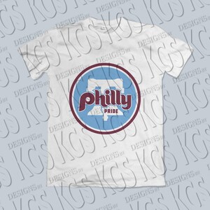 Pode incluir: Camiseta branca com um design circular. O design apresenta a palavra "Philly" em escrita marrom, sobre um fundo azul claro, com uma borda marrom. As palavras "Pride" e um "P" estilizado tamb&eacute;m est&atilde;o inclu&iacute;das.