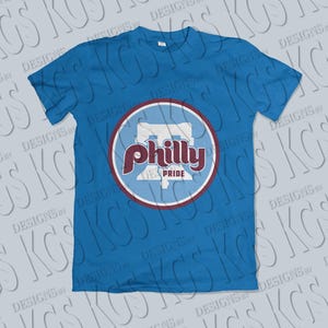 Pode incluir: Uma camiseta azul com um design circular. O design inclui as palavras "Philly Pride" em bord&ocirc; e branco, e um contorno estilizado da Pensilv&acirc;nia. A camiseta &eacute; de cor azul lisa.
