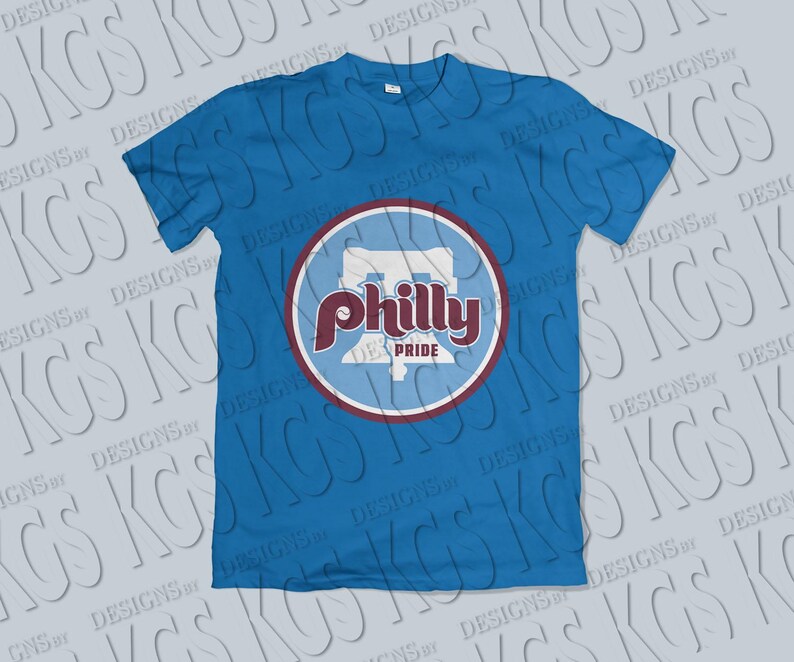 Pode incluir: Uma camiseta azul com um design circular. O design inclui as palavras "Philly Pride" em bord&ocirc; e branco, e um contorno estilizado da Pensilv&acirc;nia. A camiseta &eacute; de cor azul lisa.