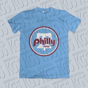Pode incluir: Camiseta azul claro com um design gr&aacute;fico circular. O design gr&aacute;fico apresenta a palavra "Philly" em escrita bord&ocirc; e as palavras "Pride" abaixo. O design gr&aacute;fico tamb&eacute;m inclui um contorno branco e uma borda bord&ocirc;. A camiseta &eacute; de gola redonda.