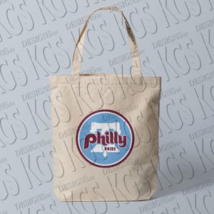 Pode incluir: Bolsa tote de lona bege com um design circular. O design inclui a palavra "Philly" em escrita marrom, um fundo azul e a palavra "Pride" em branco. A bolsa tem duas al&ccedil;as.