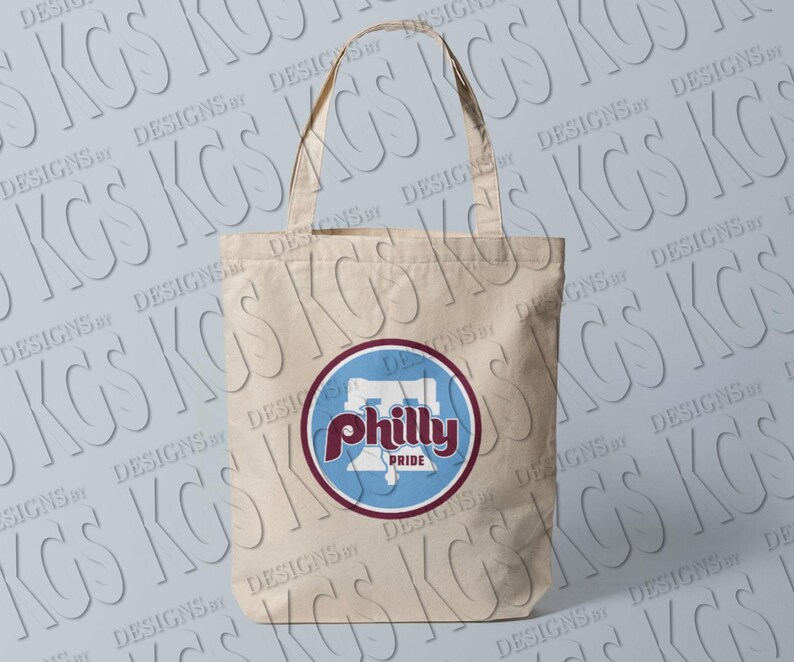Pode incluir: Bolsa tote de lona bege com um design circular. O design inclui a palavra "Philly" em escrita marrom, um fundo azul e a palavra "Pride" em branco. A bolsa tem duas al&ccedil;as.