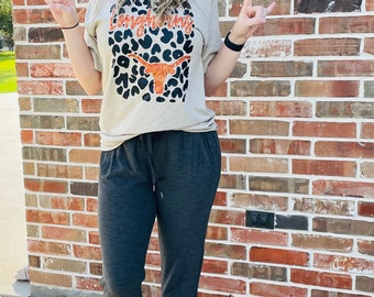 May include: A woman wearing a beige t-shirt with a black and orange leopard print and the text "Longhorns" in orange glitter. She is wearing black joggers and is standing on a rock with a background of a brick wall.