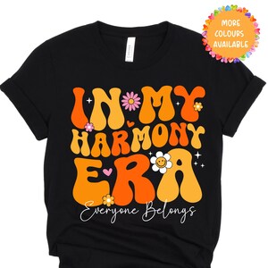 May include: Black t-shirt featuring the phrase "IN MY HARMONY ERA" in a retro orange and yellow font, with flower and heart graphics. The words "Everyone Belongs" are in white script. A colourful circle with "MORE COLOURS AVAILABLE" is in the top right corner.