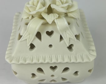 May include: White porcelain trinket box with a square shape and a delicate, openwork design. The lid features two sculpted white roses and a ribbon.