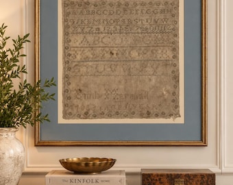 May include: A framed antique needlepoint sampler featuring alphabet letters, numbers, and the name Emb Marshall 1853, displayed in a gold frame with a blue mat. The sampler is on aged, light brown fabric with a decorative floral border pattern.