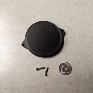 May include: A black, circular component with a textured surface, accompanied by two silver screws and a small, circular metal piece with a central hole and two smaller holes. The items are arranged on a light gray surface.