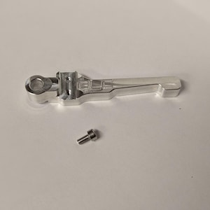 May include: A silver-coloured metal lever component featuring the engraved text GLC on its side, with a circular mounting hole at one end and a small, separate metal screw positioned directly below the main lever piece.