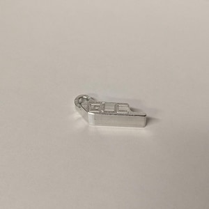 May include: A small, silver-colored metal charm featuring a rectangular body with a circular loop at one end. The letters GLC are embossed in a block font on the top surface of the charm, which has a smooth, metallic finish.