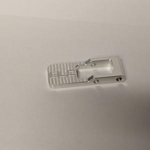 May include: A silver-colored metal component with a rectangular base featuring a striped texture and the letters GLC engraved on the surface. The piece includes a hollow rectangular cutout and two small circular holes along the side edge.