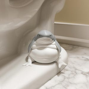 Puede incluir: Una escultura de cerámica de una criatura parecida a una rana con cabeza gris y cuerpo blanco, posada sobre una superficie blanca. La escultura tiene un diseño de dibujos animados y está cerca de un inodoro blanco.