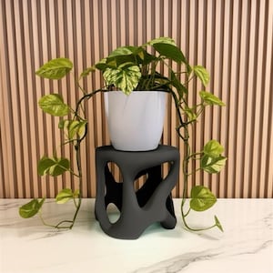 May include: A white plant pot with green and yellow variegated leaves sits on a dark grey, open-design plant stand. The plant stand is on a white countertop. The background is a wood-panelled wall.