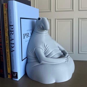 Può includere: Una scultura grigio chiaro di una creatura rilassata e seduta con un grande naso e le braccia incrociate, usata come fermalibri. È posizionata accanto a una pila di libri, tra cui uno con la parola "PRADA" sul dorso.