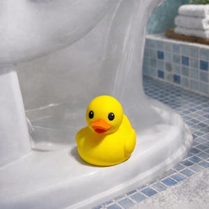 以下が含まれることがあります： 鮮やかな黄色のラバーダックで、明るいオレンジ色のくちばしと大きな黒い目をしています。アヒルは滑らかで丸みを帯びた体をしており、白い表面の上に置かれています。頭、翼、尾がはっきりと定義された、古典的なアヒルの形をしています。