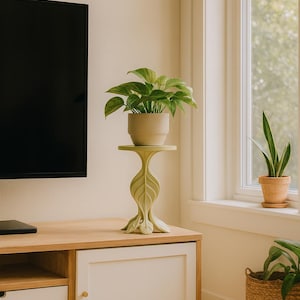 Puede incluir: Un soporte para plantas verde claro con un diseño en forma de hoja que sostiene una planta en maceta con hojas verdes. El soporte está sobre un mueble de madera con una puerta y un cajón blancos. Un televisor está en la pared.