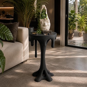 May include: A matte black side table with a unique melting drip design on the circular top and a wide, stable base. On the table sits a white ceramic planter shaped like a human face with hands covering the eyes, containing a green trailing plant.