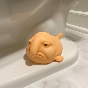 Könnte beinhalten: Pfirsichfarbene Blobfisch-Figur mit grimmigem Gesichtsausdruck. Der Fisch hat eine große Nase, kleine Augen und Flossen. Er sitzt auf einer weißen Oberfläche, mit einer weißen Toilette im Hintergrund. Die Figur ist ein dekoratives Objekt.