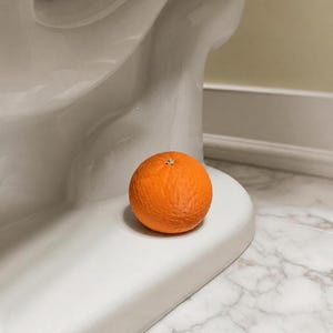 May include: A single, round, bright orange citrus fruit with a textured peel resting on the smooth, white, curved base of a porcelain toilet fixture. The fruit is positioned centrally, showcasing its vibrant colour against the white surface.
