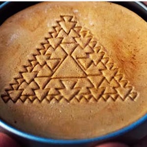 Peut inclure: Un bonbon dalgona rond, brun clair, avec un motif triangulaire géométrique pressé sur sa surface. Le bonbon est dans une petite boîte ronde de couleur argentée avec un bord bleu. La surface du bonbon est légèrement inégale.