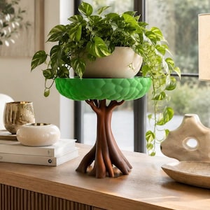 May include: A decorative plant stand shaped like a tree with a brown trunk and a bright green, textured, circular top. A white ceramic bowl containing a trailing green and white pothos plant sits securely on the flat, circular green platform.