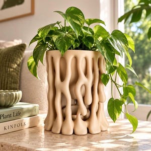 May include: A beige cylindrical plant pot featuring a decorative exterior layer of vertical, organic, drip-like structures. A green and yellow variegated pothos plant grows from the top of the pot, with leaves cascading over the sides.