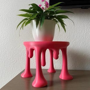 May include: A vibrant pink plant stand with a unique, melted design. The stand supports a white pot with a green plant and pink flowers. The stand's legs have a dripping effect, adding a modern touch to the decor.