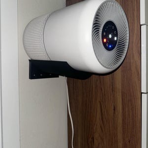 Op de afbeelding: Een witte luchtreiniger met een zwarte wandbeugel. De luchtreiniger heeft een ronde voorkant met een zilveren rooster en een bedieningspaneel met lampjes. De luchtreiniger is gemonteerd op een houten oppervlak.