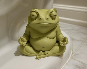 Puede incluir: Figura de rana verde claro en posición de yoga sentado. La rana tiene una expresión relajada y está colocada sobre una superficie blanca. La figura es un elemento decorativo caprichoso.