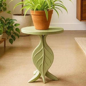 May include: A pale green side table with a leaf-shaped base and a circular top. A terracotta pot with a green plant is on the table. The table is a decorative piece for indoor plants, approximately 40 cm in height.