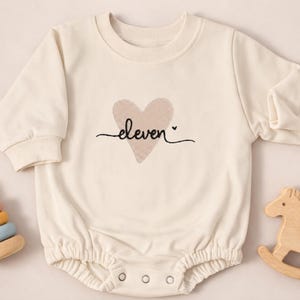 Op de afbeelding: Crèmekleurige babyromper met lange mouwen en een ronde halslijn. Het heeft een getextureerd hart en het woord "eleven" in script. Een houten schommelpaard en een stapel kleurrijke ringen zijn zichtbaar.