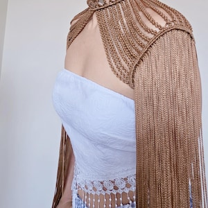 Op de afbeelding: Een metallic bronskleurig macramé schouderstuk met een hoge choker-kraag voorzien van reflecterende spiegelaccenten, ingewikkelde gevlochten koorddetails over de borst en lange, vloeiende franjes die langs de armen naar beneden vallen.