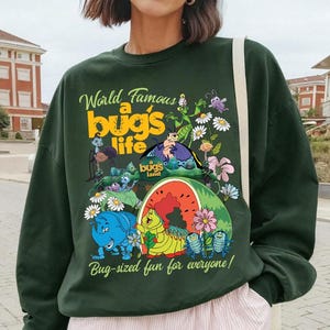 May include: Dark green sweatshirt featuring a colorful graphic from "a bug's life" with the text "World Famous a bug's life" and "Bug-sized fun for everyone!". The design includes cartoon bugs, flowers, and a watermelon.