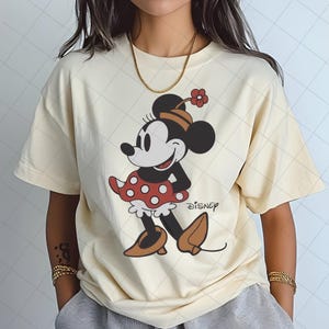 May include: Cream-colored t-shirt featuring a cartoon illustration of Minnie Mouse. Minnie is depicted in a red polka-dot dress, brown shoes, and a hat with a flower. The Disney logo is printed below the character.