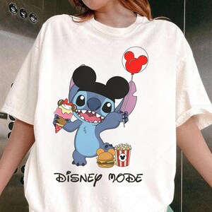 May include: Off-white t-shirt featuring a blue cartoon character with large ears, holding a red Mickey Mouse balloon and an ice cream cone. The character is also wearing a black hat. The shirt has the words "DISNEY MODE" printed in black.