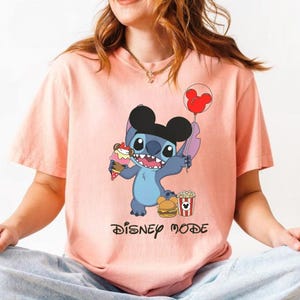 May include: A light pink t-shirt featuring a cartoon character holding ice cream, a balloon, a burger, and popcorn. The character is blue with black ears. The text "DISNEY MODE" is printed below the character.