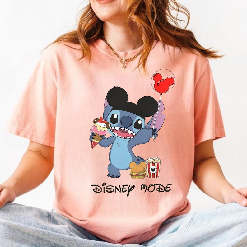 May include: A light pink t-shirt featuring a cartoon character holding ice cream, a balloon, a burger, and popcorn. The character is blue with black ears. The text "DISNEY MODE" is printed below the character.
