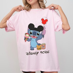 May include: A light pink t-shirt featuring a cartoon character holding an ice cream cone and a Mickey Mouse balloon. The character is wearing a black hat and is surrounded by popcorn and a burger. The words "DISNEY MODE" are printed below.