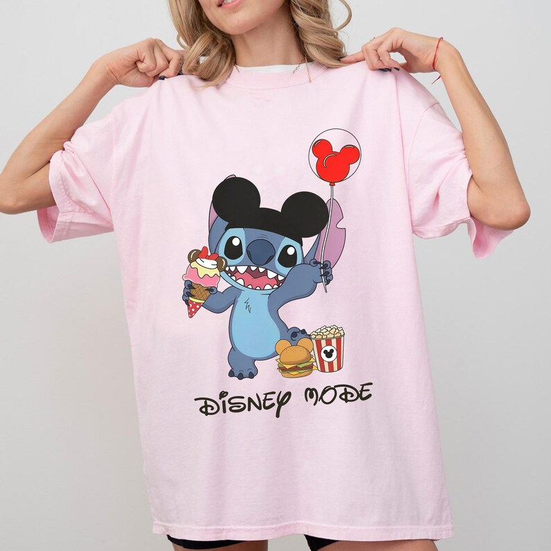 May include: A light pink t-shirt featuring a cartoon character holding an ice cream cone and a Mickey Mouse balloon. The character is wearing a black hat and is surrounded by popcorn and a burger. The words "DISNEY MODE" are printed below.