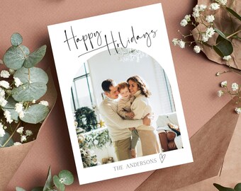 Può includere: Un biglietto bianco con il testo "Happy Holidays" in corsivo nero. Una foto di una famiglia di tre persone è al centro del biglietto. La famiglia è in piedi davanti a un albero di Natale. Il testo "The Andersons" è in fondo al biglietto.