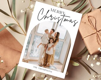 Può includere: Un biglietto di Natale bianco con la scritta "MERRY CHRISTMAS" in elegante corsivo. Il biglietto presenta una foto di famiglia all'interno di un arco, con il cognome in basso. Un regalo confezionato e accenti floreali completano la scena festosa.