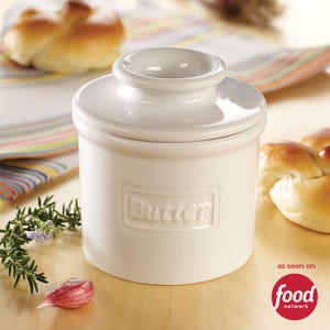Op de afbeelding: Een witte keramische botervloot met een deksel. Het woord "Butter" is in reliëf op de voorkant. De schaal is cilindrisch met een kleine, ronde handgreep op het deksel. Het Food Network-logo staat rechtsonder.