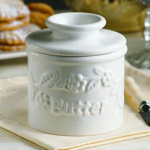 May include: A white ceramic butter dish with a lid. The dish has embossed floral detailing and the word "Butter" on the front. A butter knife rests on a yellow napkin next to the dish. The background includes baked goods.