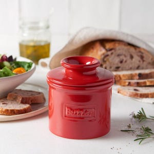 Puede incluir: Un mantequero de cerámica rojo brillante con tapa, con la palabra "Butter" en relieve. El plato cilíndrico tiene una parte superior redondeada. La imagen muestra una mesa con pan, ensalada y aceite de oliva.