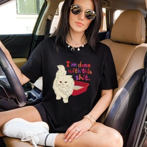 Puede incluir: Camiseta negra con el texto "I'm done with this shit" y una ilustración de gato de dibujos animados. El gato es de color crema y la caja de arena es roja. La persona lleva gafas de sol y está sentada en un coche.