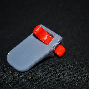 May include: A small, gray 3D-printed object with two bright red, rectangular components. The object has a curved, rectangular base and a slot to hold the red pieces. The item is set against a dark background.