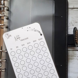 May include: A white card with a star and shooting star design. The card has a grid of circles with dollar amounts listed at the top: $1, $5, $10, $20, $50, and $100. The card is labeled "Star Note Tracker".